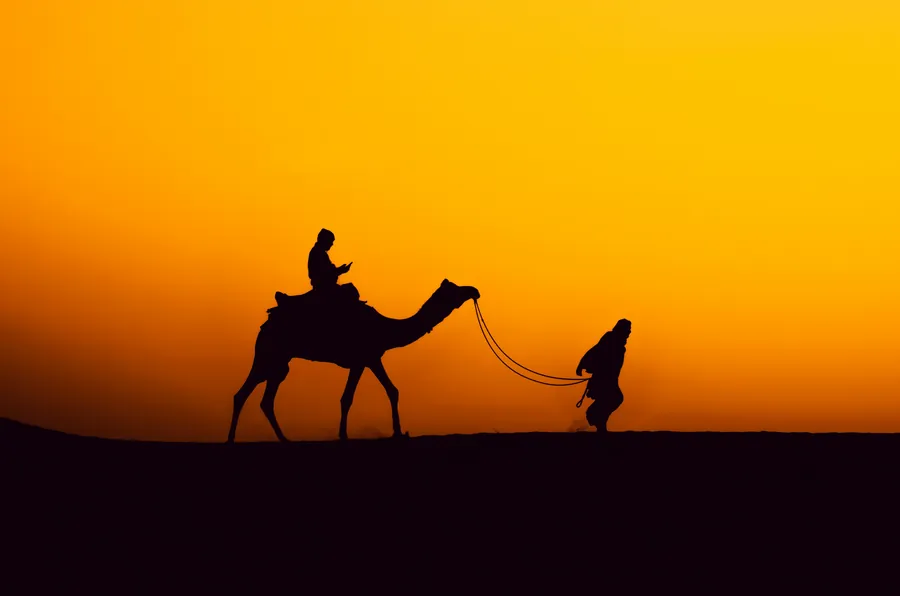 A camel and its handler silhouetted against an orange desert sunset